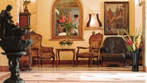 A lobby with a statue, two armchairs, a table with a vase of flowers, a lamp, and a framed painting. - Hotel Europejski (Kraków)
