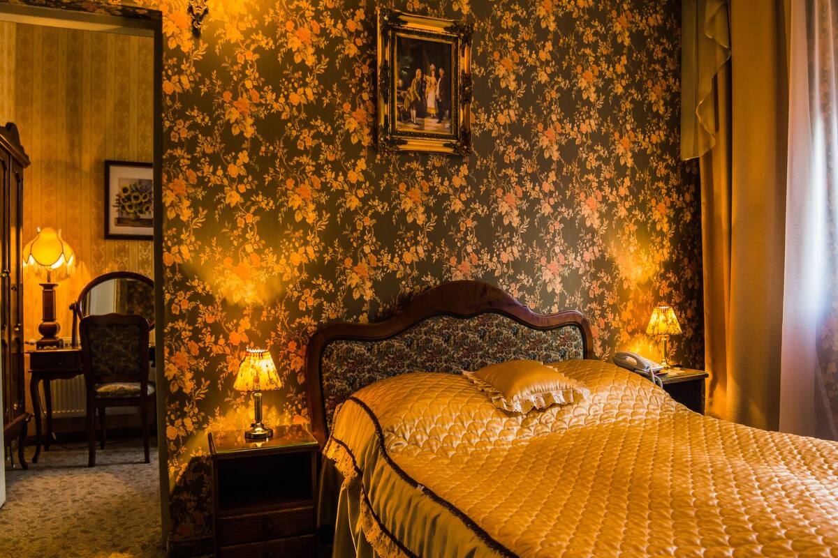 A bedroom with a floral wallpaper, a wooden headboard, a bed with a quilt, two bedside lamps, a nightstand, a chair, and a framed picture on the wall.