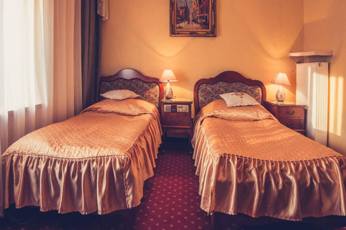 A hotel room with two beds, a nightstand with a lamp, a painting on the wall, and a window with curtains.