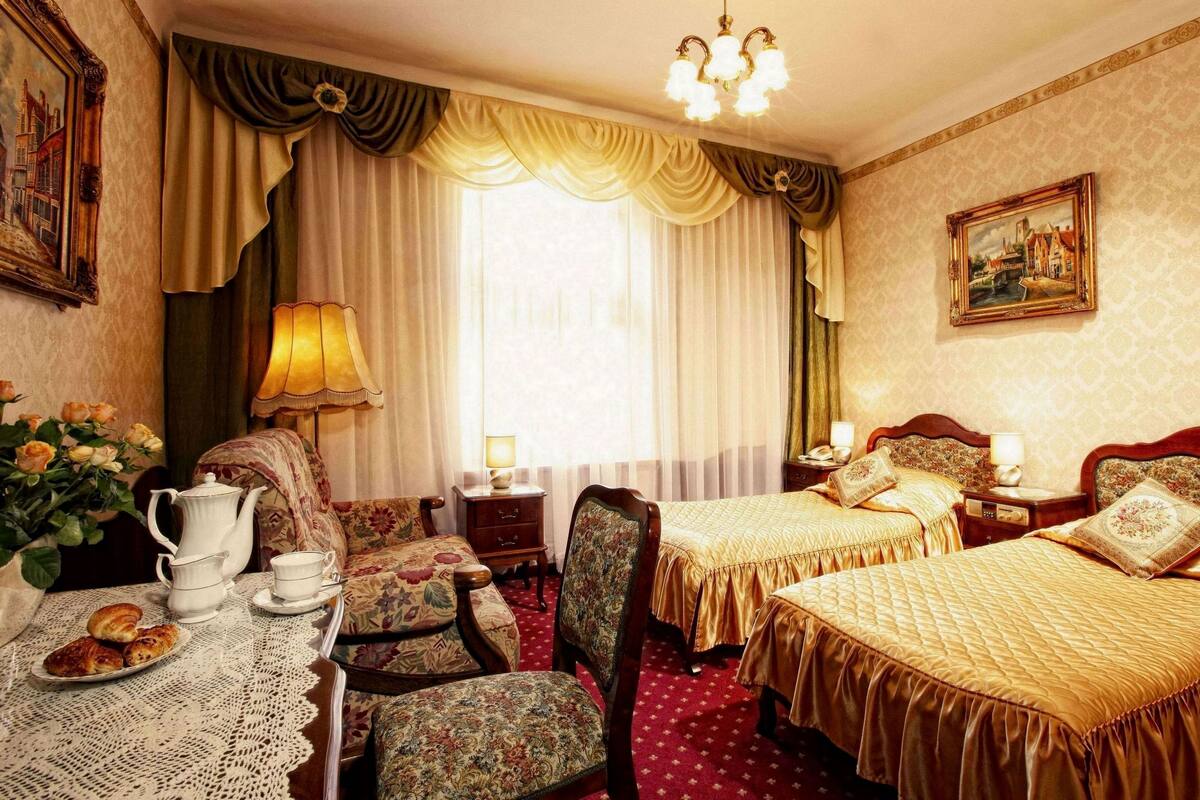 A hotel room with two beds, a dining table with a pitcher and teapot, floral armchairs, a chandelier, and a painting on the wall.