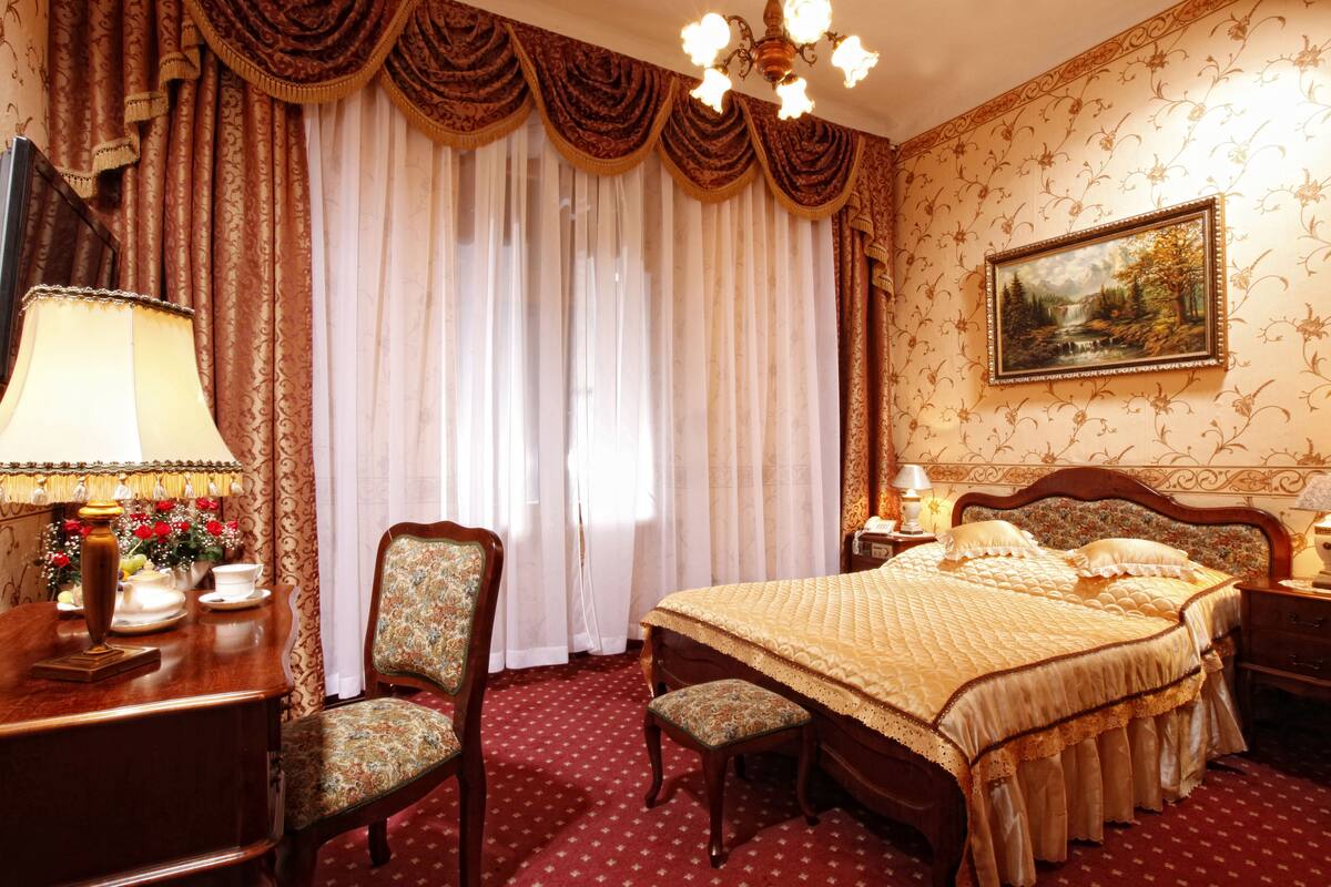 A traditional bedroom with a large bed, a wooden nightstand, a desk with a lamp, a chair, and curtains.
