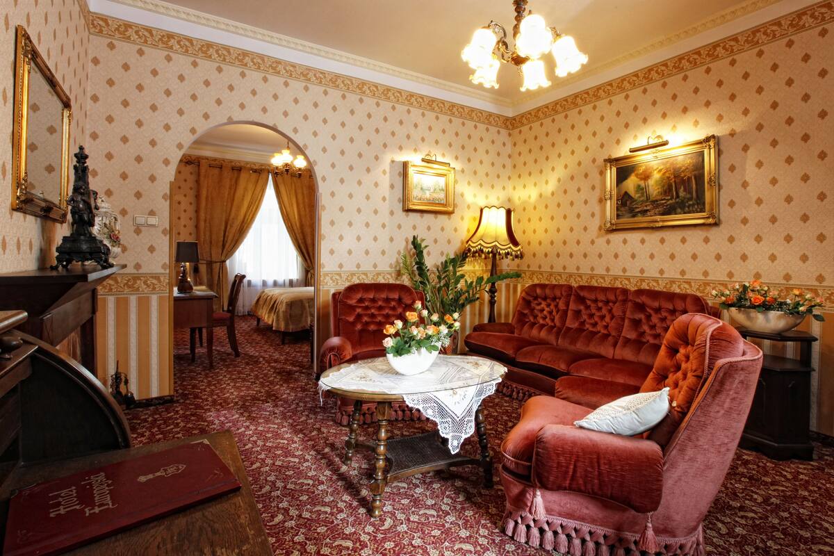 A room with a red velvet sofa, a wooden coffee table with a lace tablecloth, a red armchair, a wooden side table with a vase of flowers, a chandelier, a bed with a white canopy, and a mirror with a statue.