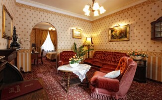 Una habitación con un sofá de terciopelo rojo, una mesa de centro de madera con mantel de encaje, un sillón rojo, una mesita auxiliar de madera con un jarrón de flores, una araña, una cama con dosel blanco y un espejo con una estatua.
