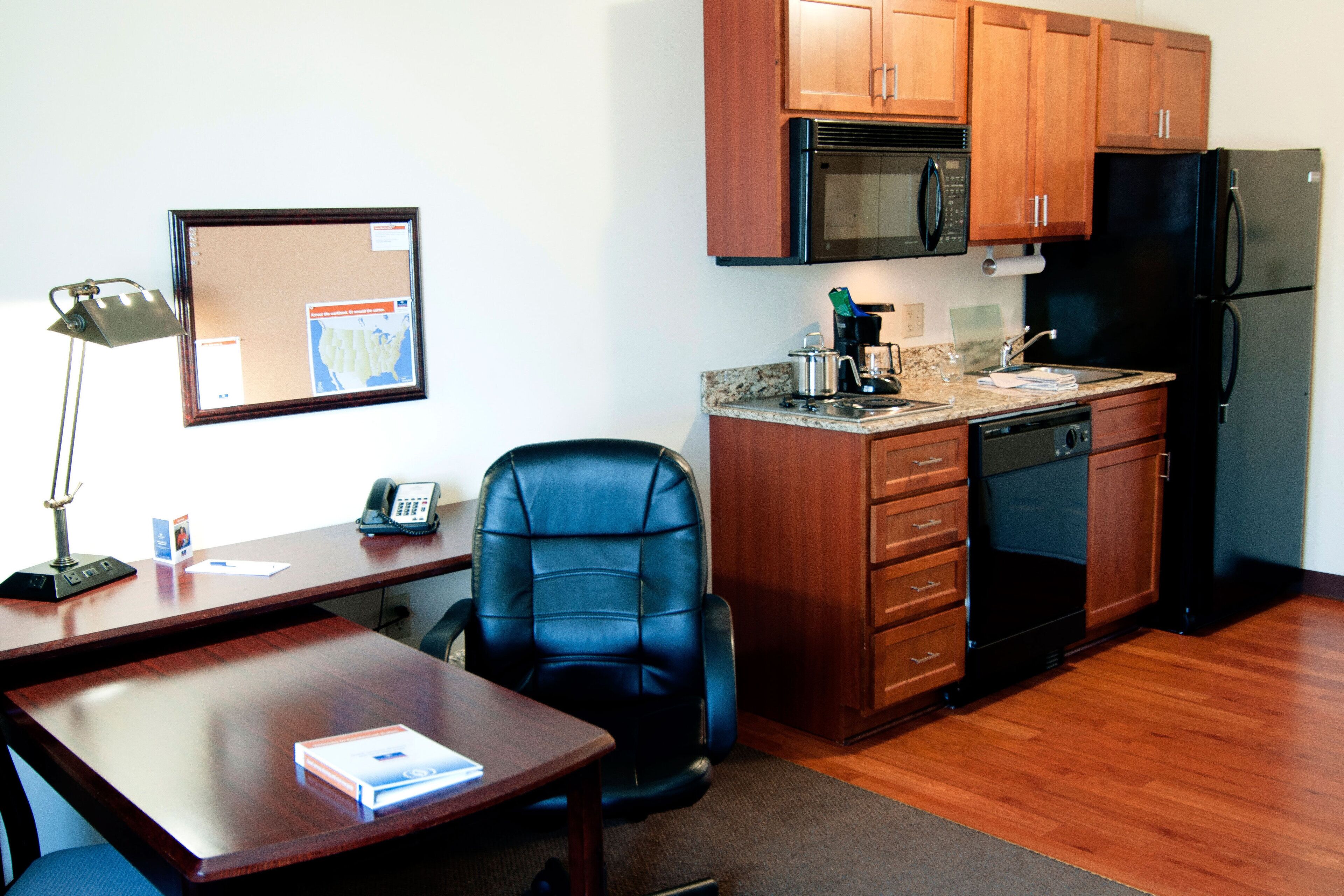 A compact kitchen with wooden cabinets, a microwave, a refrigerator, a coffee maker, and a desk with a chair.