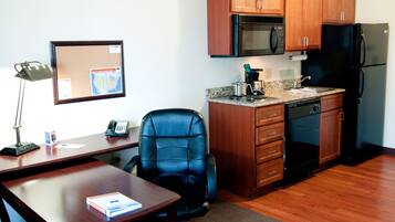 A compact kitchen with wooden cabinets, a microwave, a refrigerator, a coffee maker, and a desk with a chair.