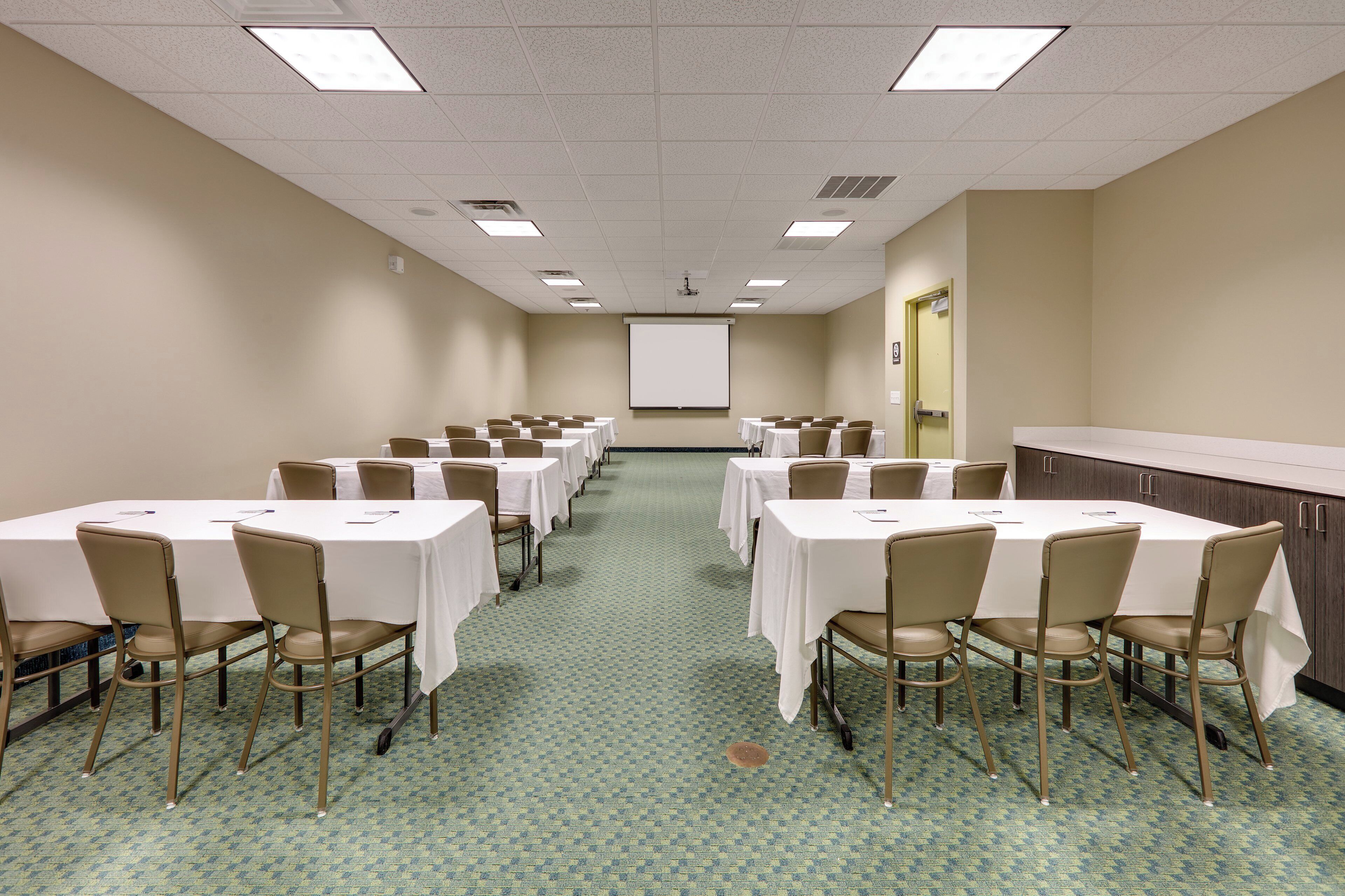 Una sala conferenze con diversi tavoli e sedie, uno schermo per proiettore e un pavimento con tappeto.