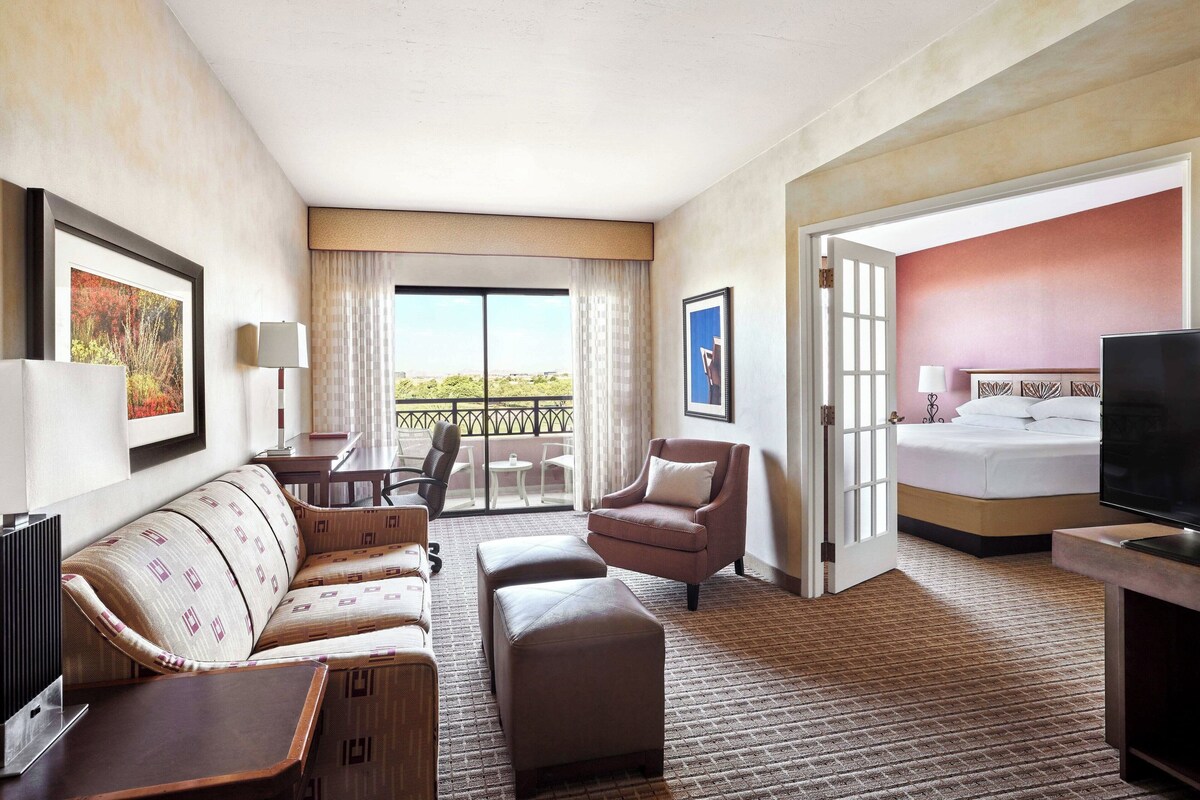 Een hotelkamer met een bank, fauteuil, poef, een televisie, een bed en een balkon met uitzicht.