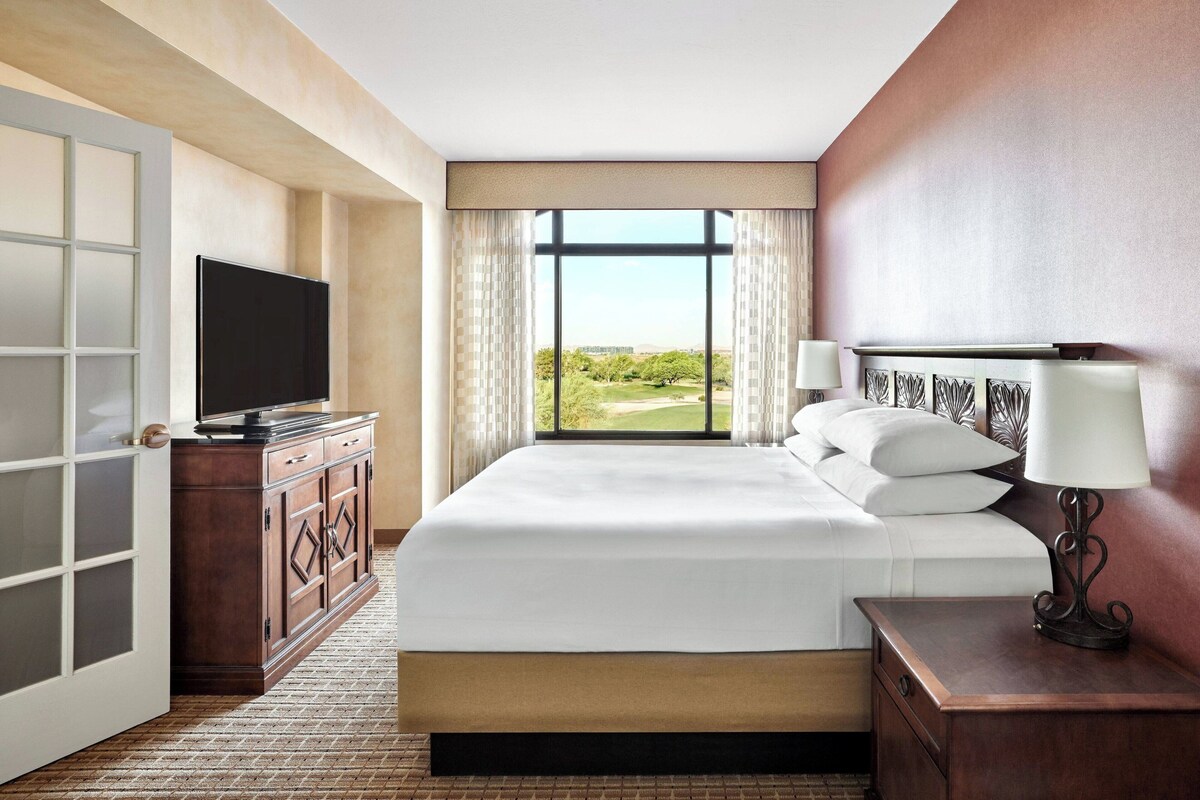 Een hotelkamer met een groot bed, een televisie op een houten kast, uitzicht op een groen landschap door het raam en een nachtkastje met een lamp.