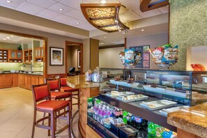 Zona de desayuno de hotel con mostrador, asientos y una vitrina con alimentos.