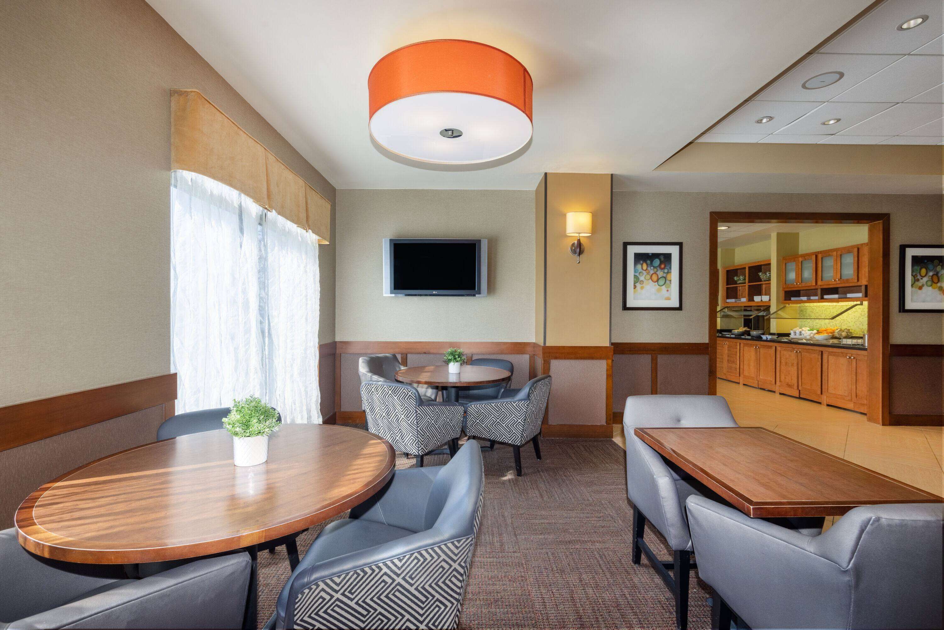 A hotel dining area with a round table, chairs, a television, and a kitchen in the background.
