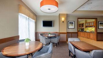 A hotel dining area with a round table, chairs, a television, and a kitchen in the background.
