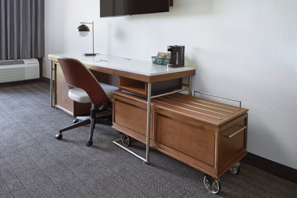 A compact hotel room with a wooden desk, a modern chair, a small cart, and a flat-screen TV mounted on the wall.