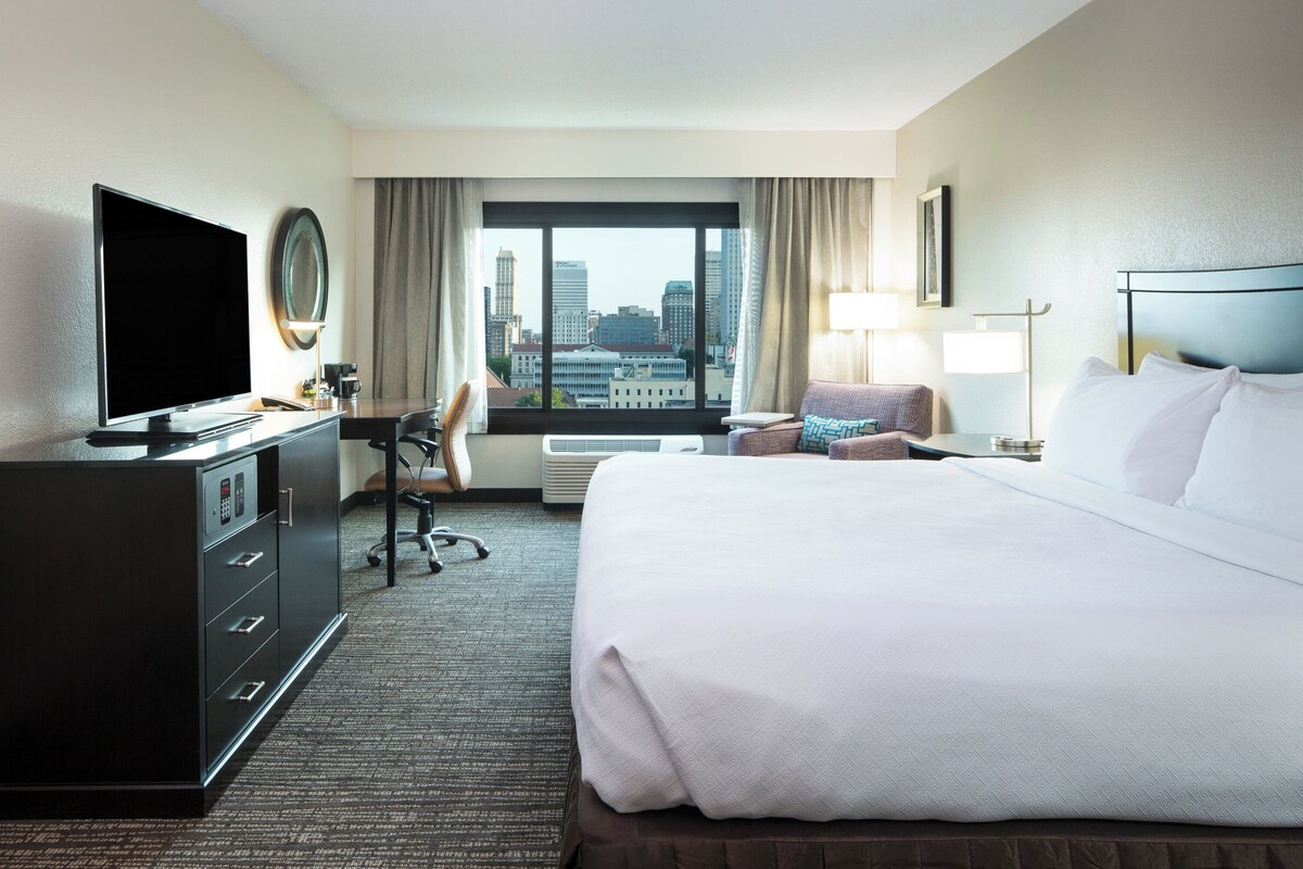 Une chambre d’hôtel avec un grand lit, un bureau avec un ordinateur, une télévision et une vue sur le paysage urbain.