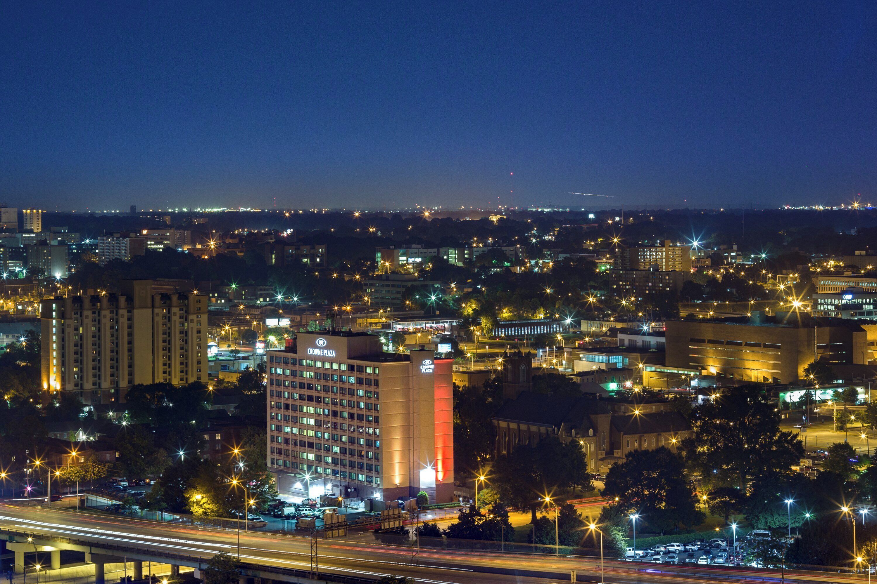 A cityscape at night with illuminated buildings, a prominent hotel named 'Crowne Plaza,' and a highway with light trails.