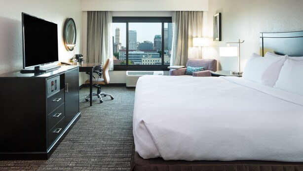 Une chambre d’hôtel avec un grand lit, un bureau avec un ordinateur, une télévision et une vue sur le panorama de la ville.