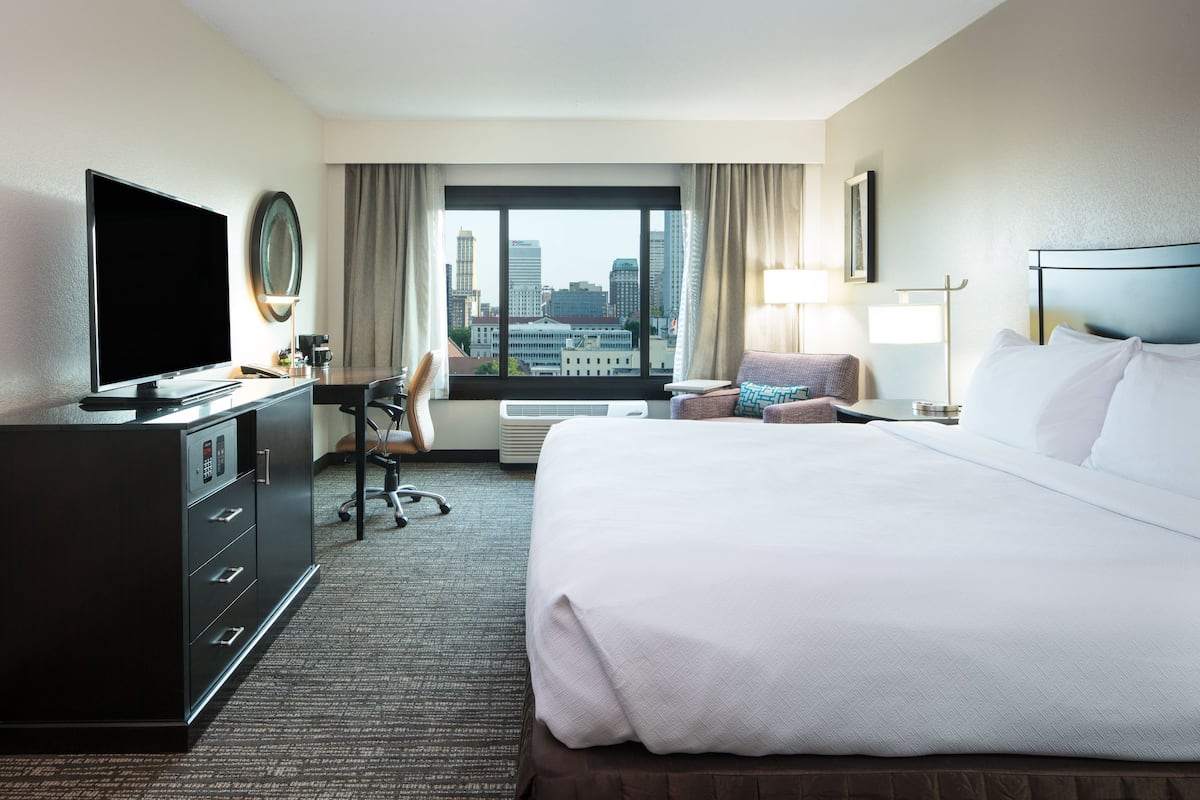 Habitación de hotel con una cama grande, un escritorio con computadora, televisor y vistas del horizonte urbano.
