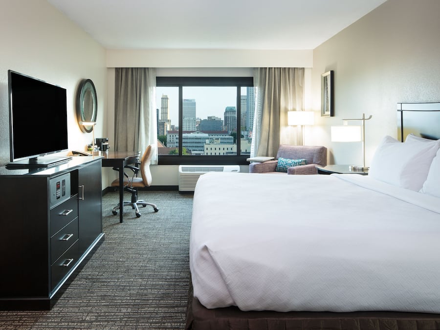 Habitación de hotel con una cama grande, un escritorio con computadora, televisor y vistas del horizonte urbano.