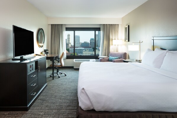 Une chambre d’hôtel avec un grand lit, un bureau avec un ordinateur, une télévision et une vue sur le panorama de la ville.