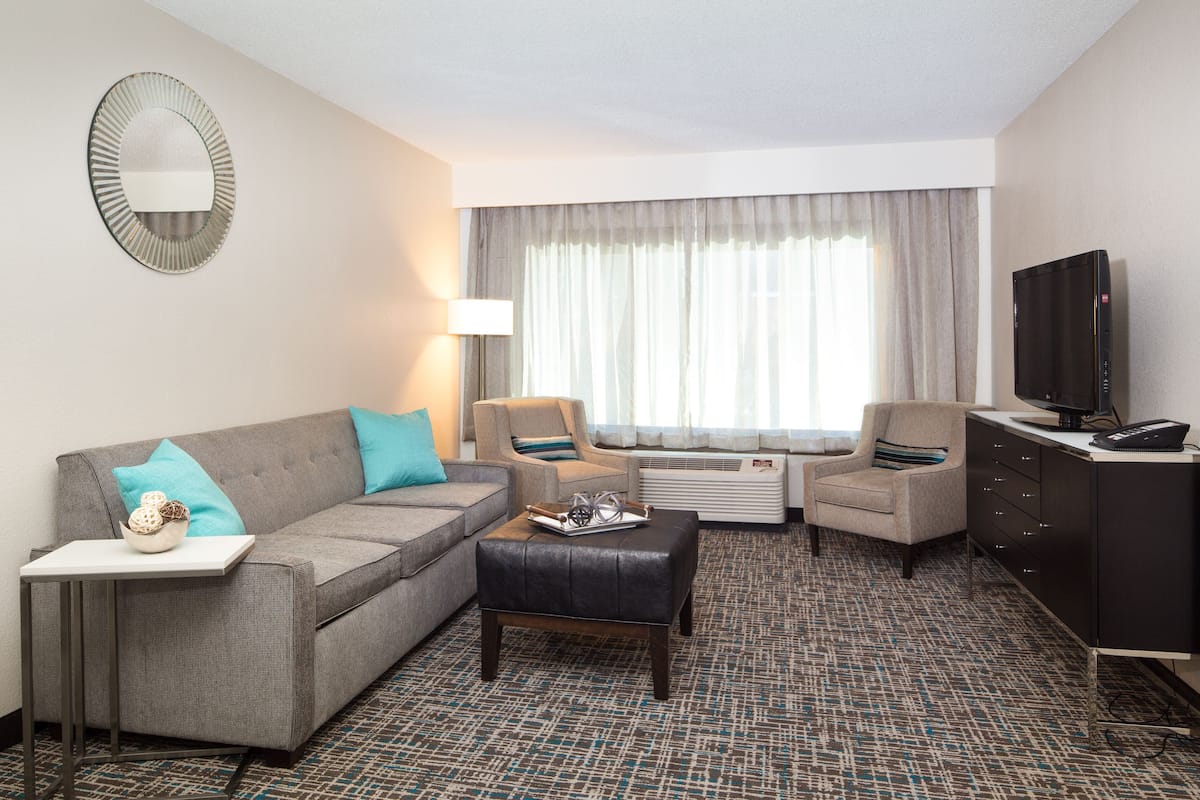Habitación de hotel con sofá gris, dos sillones, una mesa de centro, televisor y minibar.