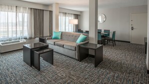 Une chambre d’hôtel avec un canapé, deux tables d’appoint, une lampe, une table à manger avec des chaises et une grande fenêtre avec des rideaux.