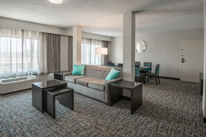 Une chambre d’hôtel avec un canapé, deux tables d’appoint, une lampe, une table à manger avec des chaises et une grande fenêtre avec des rideaux.