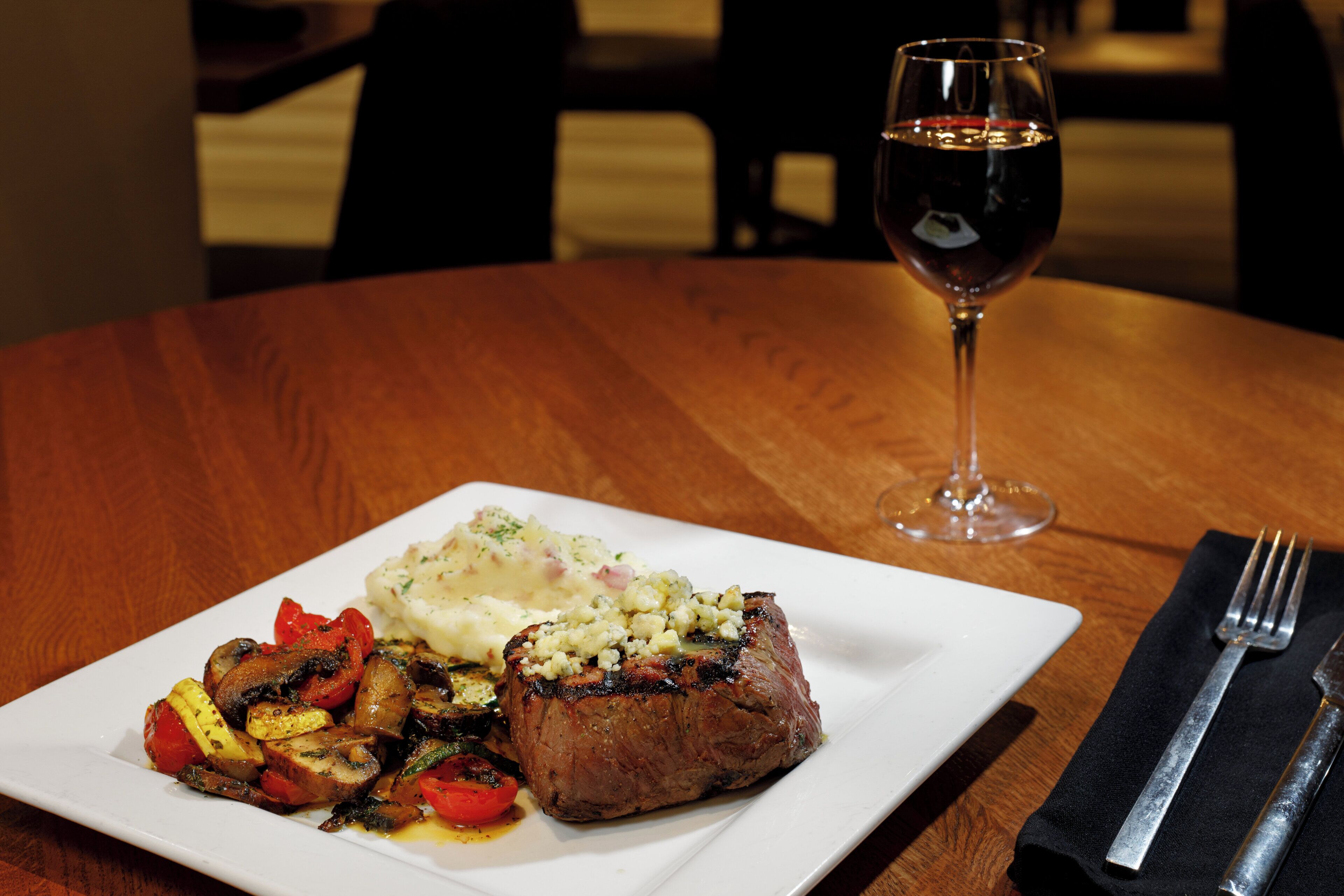 Un plato de comida con un bistec, verduras y puré de papas sobre un plato blanco, con una copa de vino en una mesa de madera.