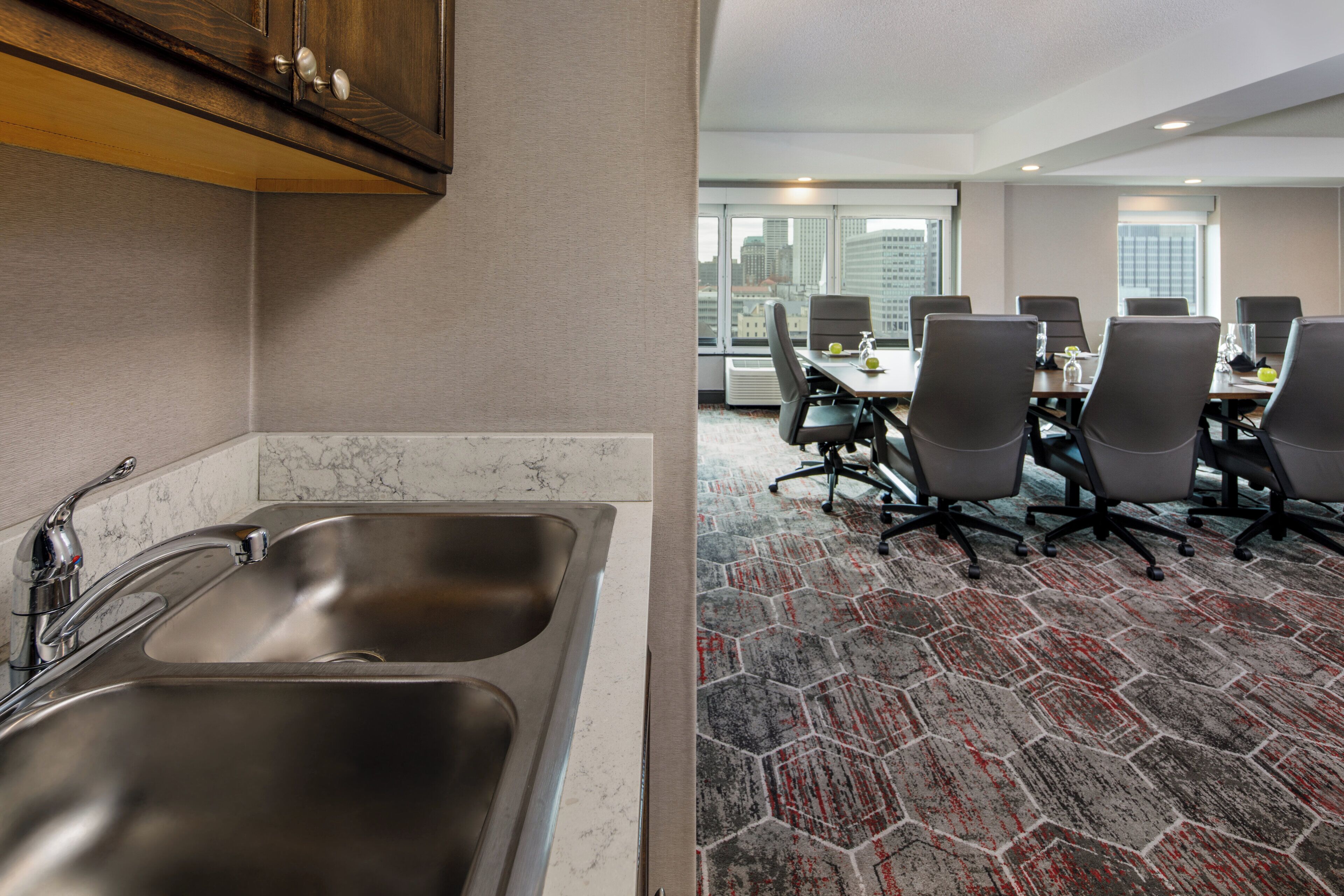 Habitación de hotel con cocina, una mesa de comedor y sillas, y vistas de la ciudad a través de amplias ventanas.