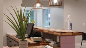 Un comptoir d’accueil orné d’une plante en pot, d’une pile de magazines, d’un flacon de gel hydroalcoolique et d’un plan de travail en bois.