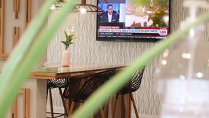 Un espace repas moderne avec une table en bois, des chaises noires et un téléviseur diffusant un journal télévisé.