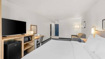 A hotel room with a bed, a desk with a television, a microwave, a chair, a sofa, and a door.