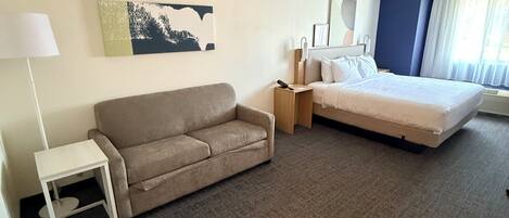 A hotel room with a sofa, a bed, a nightstand, a lamp, and a painting on the wall.