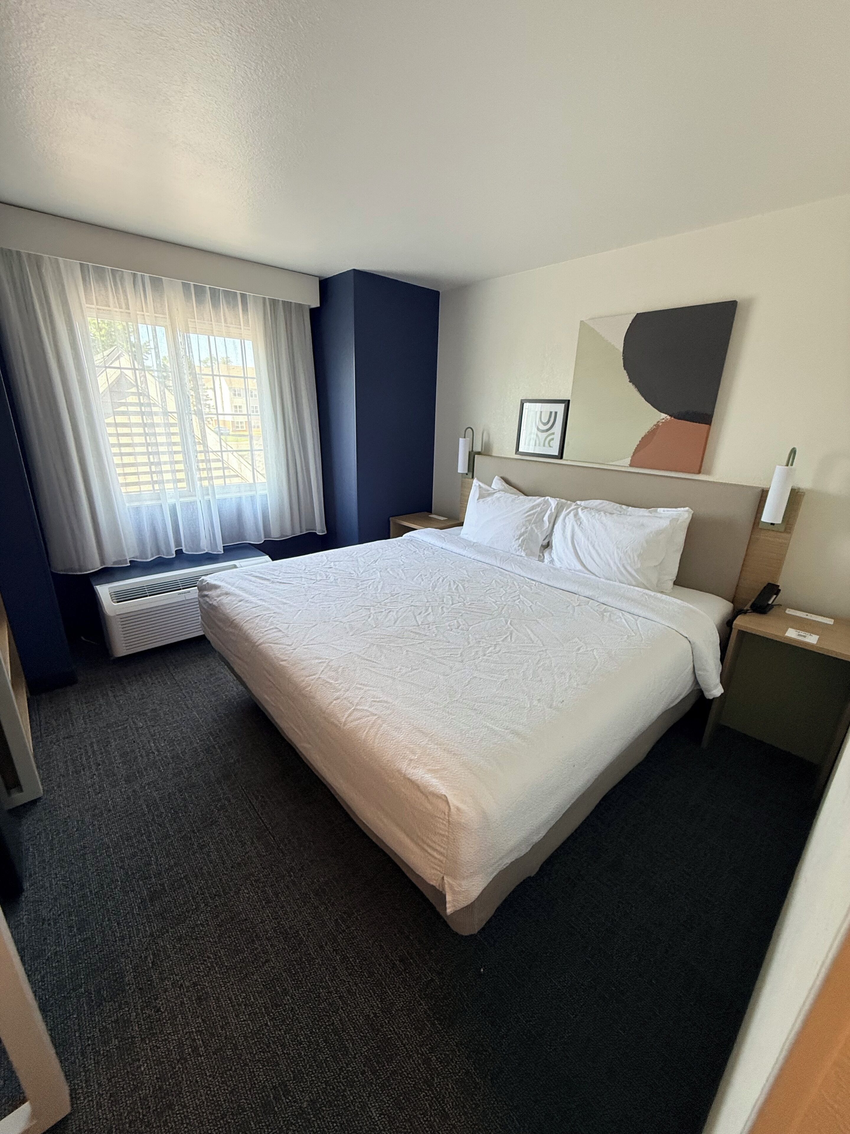 Habitación de hotel con una cama grande, una ventana con cortinas, un radiador, una mesita de noche con un teléfono y una obra de arte colgada en la pared.