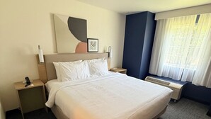 A hotel room with a large bed, a bedside table, a nightstand, a wall-mounted lamp, a window with curtains, and a partial view of a wall with a geometric design. - Spark by Hilton Appleton (Appleton)