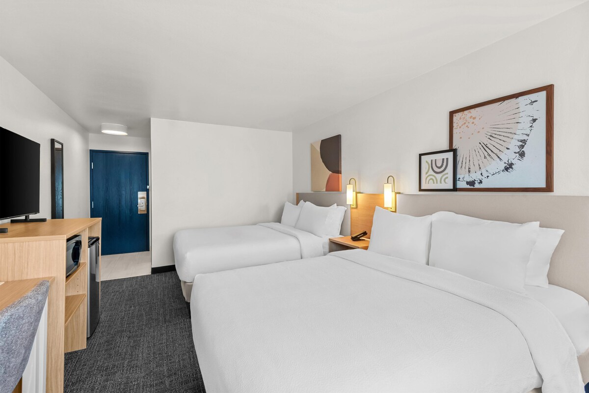 A hotel room with two beds, a desk with a flat-screen TV, a door, and artwork on the wall.