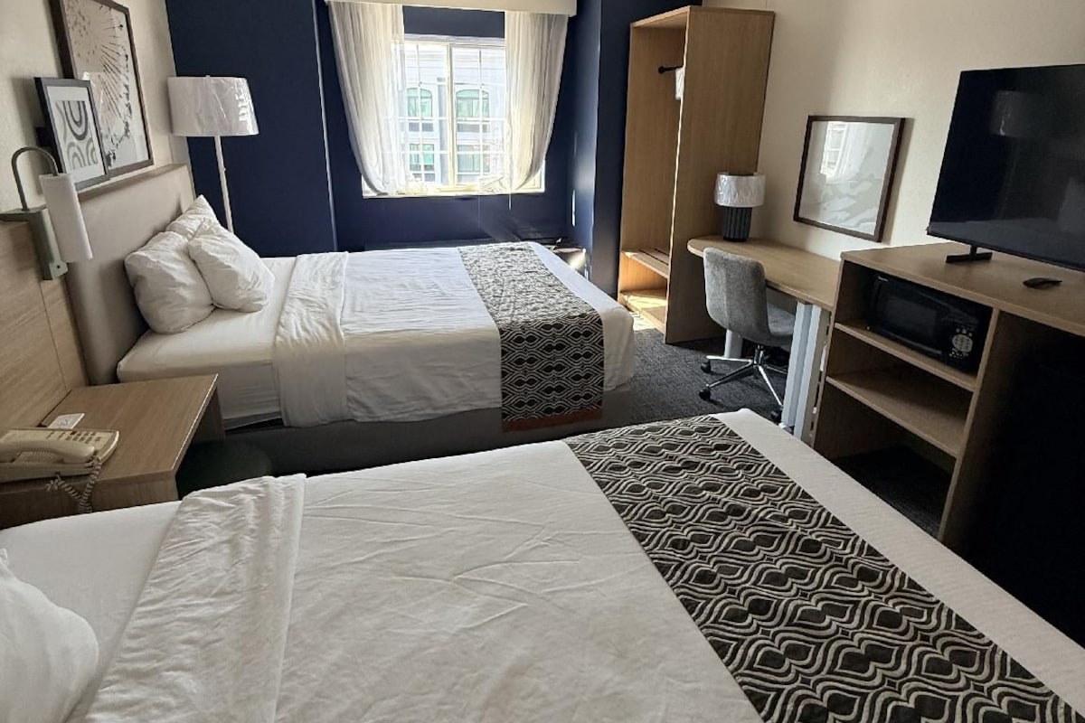 A hotel room with two beds, a desk, a television, and a window with curtains.