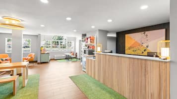 A hotel reception area with a wooden reception desk, a large abstract painting, and a view of the outdoors through large windows.