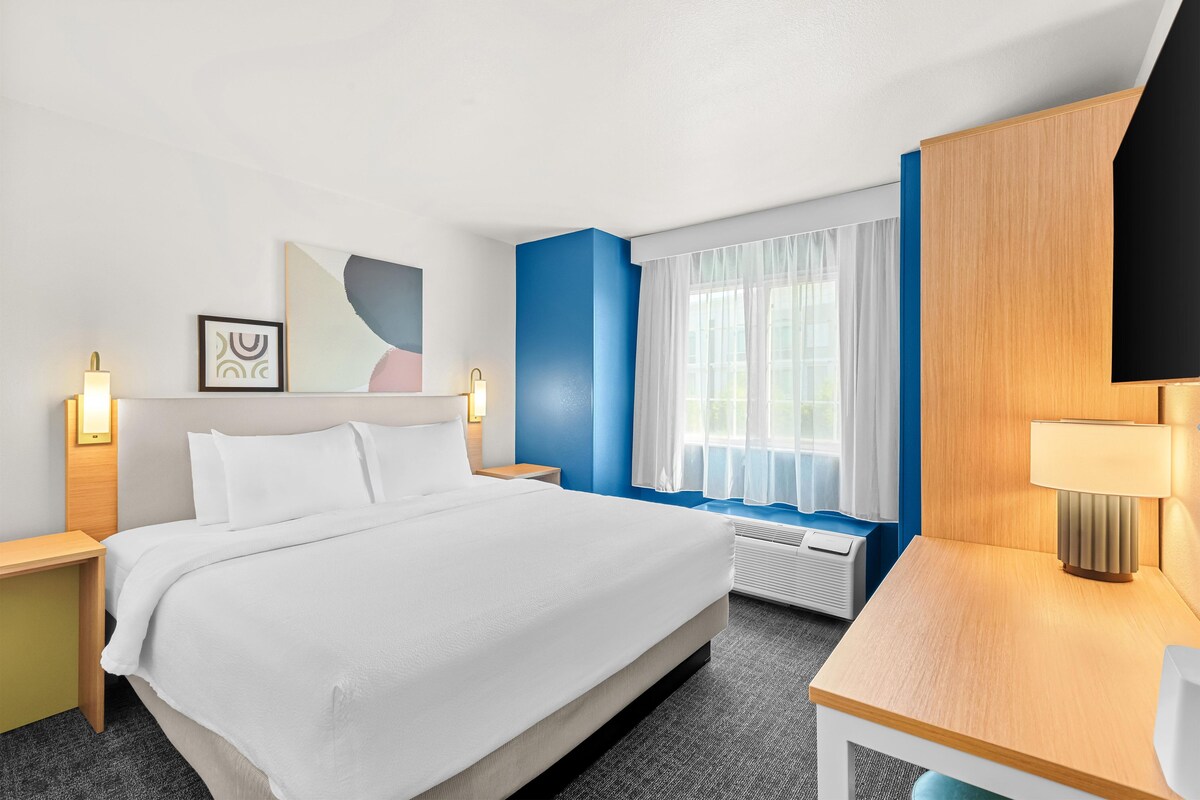 A hotel room with a large bed, bedside tables, a desk, a window with curtains, and wall art.