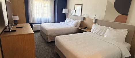 A hotel room with two beds, a desk, a chair, a TV, a lamp, and a painting on the wall.