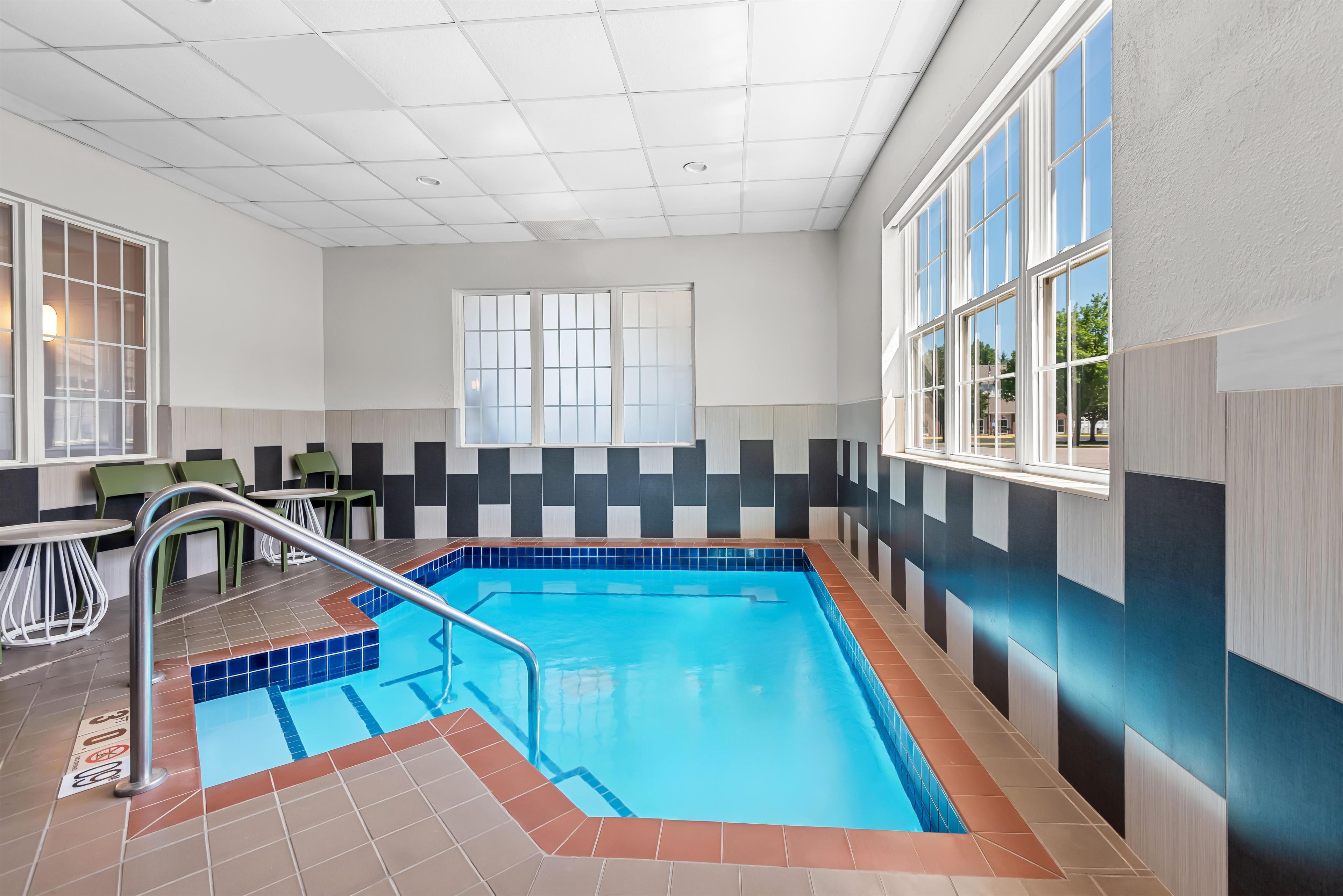 An indoor swimming pool with a tiled floor, a metal pool ladder, and a window with a view of the outside.