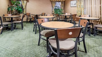 A hotel dining area with round tables and chairs, green patterned carpet, and potted plants.