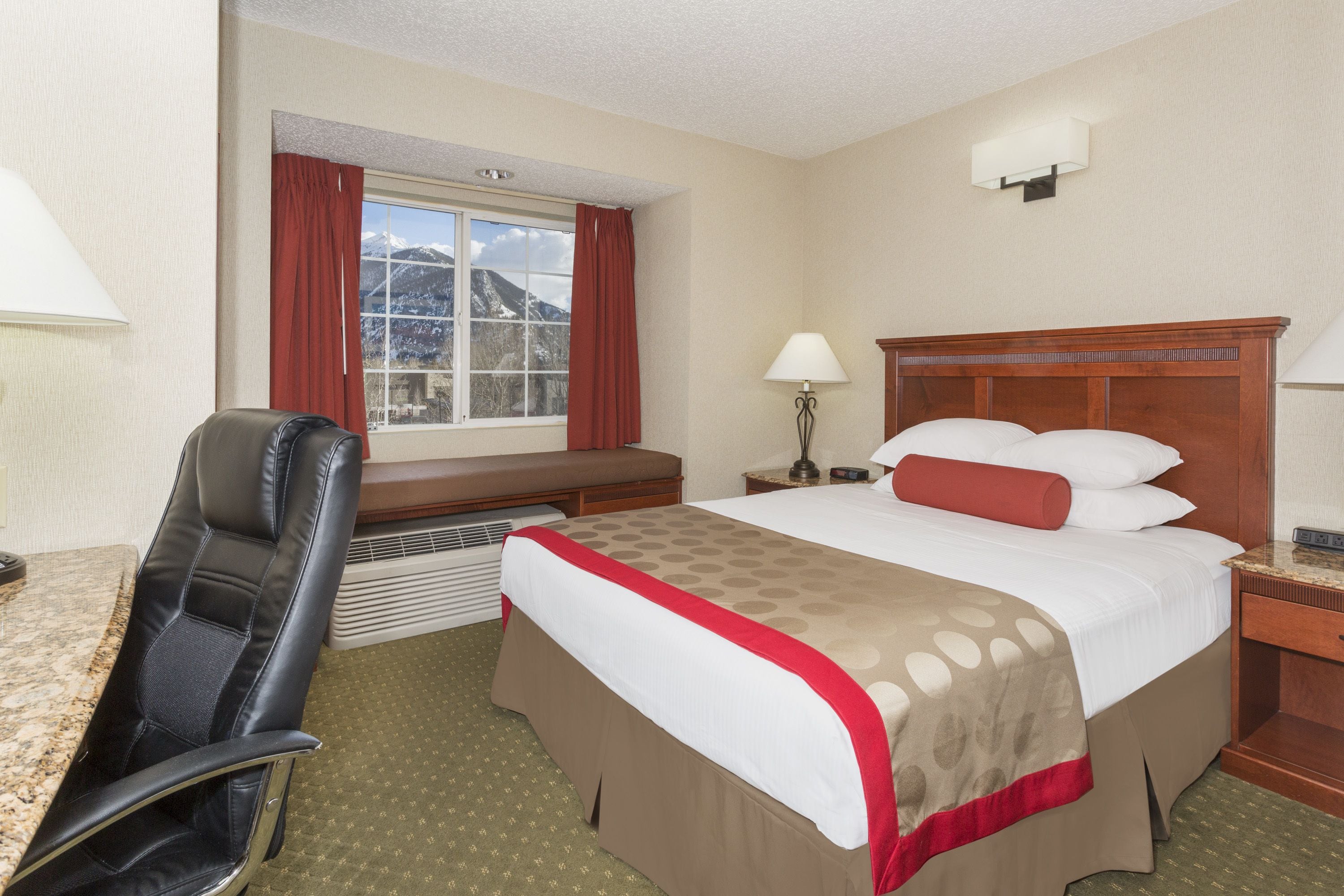 A hotel room with a bed, a desk, a chair, a window with curtains, and a lamp.