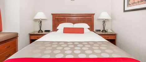 A hotel room with a wooden headboard, two bedside tables with lamps, a bed with a polka dot bedspread, and a red accent pillow.