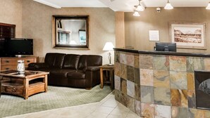 A hotel lobby with a leather sofa, a wooden coffee table, a TV, a reception desk, and a stone wall with a framed picture. - Ramada by Wyndham Frisco (Frisco)