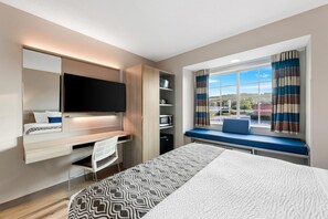 A modern hotel room with a bed, a desk, a TV, a window with curtains, and a view of the outside. - Microtel Inn & Suites by Wyndham Greenville / Woodruff Rd (Greenville)
