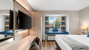 A hotel room with a bed, a desk with a chair, a television, a window with curtains, and a bedside table with a lamp. - Microtel Inn & Suites by Wyndham Greenville / Woodruff Rd (Greenville)