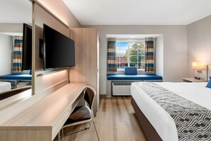 A hotel room with a bed, a desk with a chair, a television, a window with curtains, and a bedside table with a lamp. - Microtel Inn & Suites by Wyndham Greenville / Woodruff Rd (Greenville)