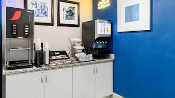 Zona de bar en un hotel, con dispensador de zumo, cuadros en la pared y una máquina de café.