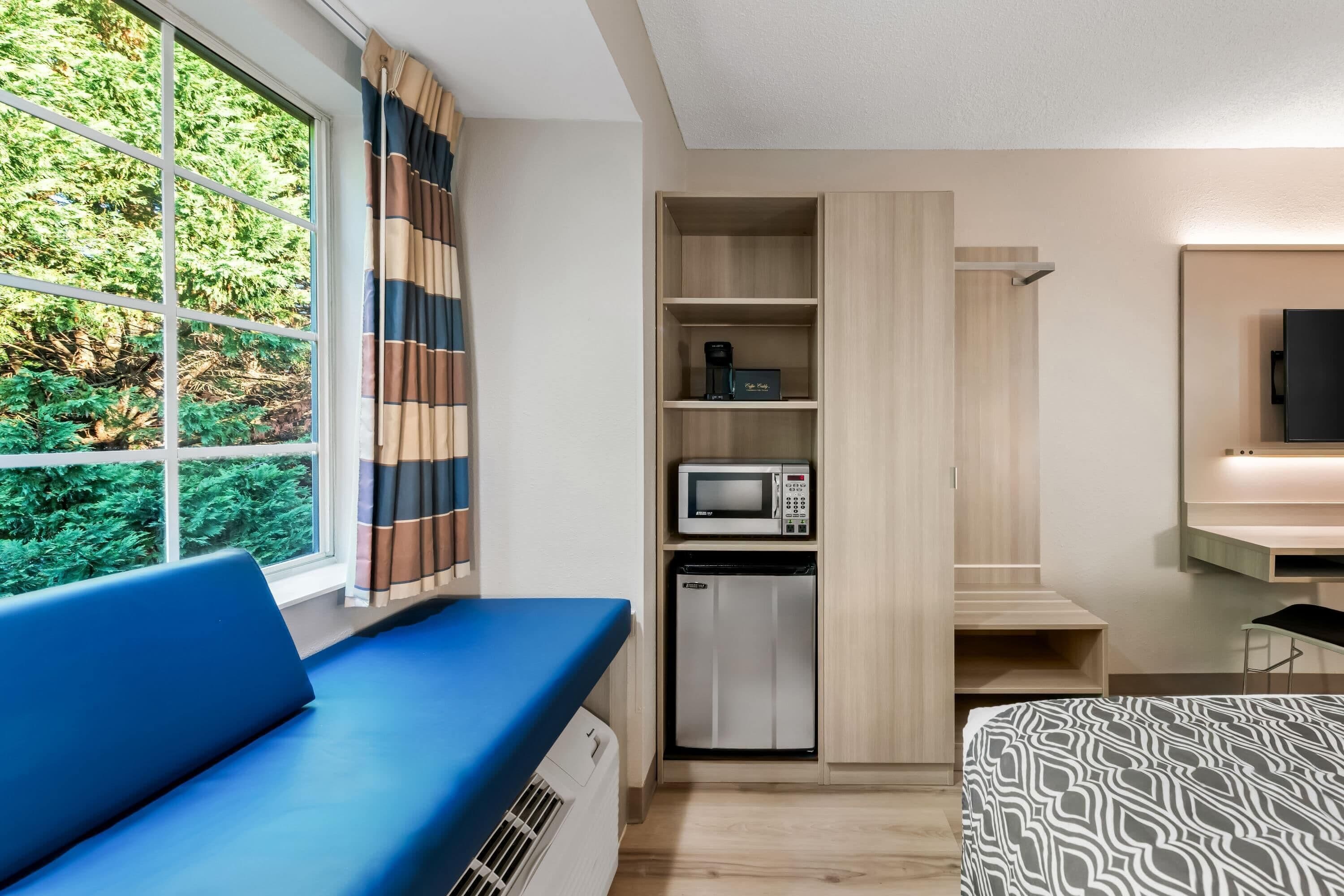 A modern hotel room with a blue sofa, a microwave, a mini fridge, and a TV mounted on the wall.
