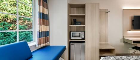 Een moderne hotelkamer met een blauwe bank, een magnetron, een minibar en een aan de muur bevestigde televisie.