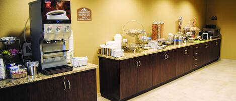 A hotel breakfast area with a coffee machine, tea station, and various food items on a counter.